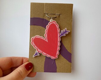 May include: A heart-shaped keychain featuring a red center with a pink scalloped border and a purple arrow piercing through it. The keychain is attached to a rectangular brown cardboard backing that has a purple painted stripe across the surface.