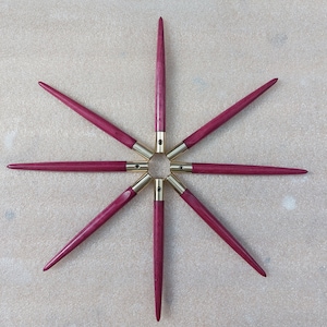 Puede incluir: Siete agujas de tejer de madera con puntas doradas dispuestas en forma de estrella sobre una superficie beige.