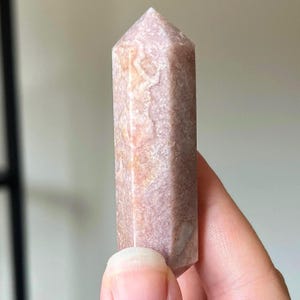 May include: A pink and tan crystal obelisk with a pointed top. The crystal has a textured surface with natural patterns and colour variations. The obelisk is held in a hand, showing its size and shape.