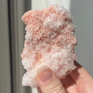 May include: A natural crystal cluster, displaying a mix of white and pale pink colours. The mineral formation has a rough, textured surface and a crystalline structure. The crystal is held in a hand.