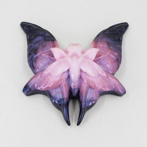 Puede incluir: Un broche de resina en forma de mariposa de color rosa y morado con un diseño detallado. Las alas son translúcidas y tienen un acabado brillante.