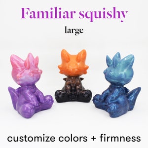 May include: Three small, colorful, hand-painted,  squishy figurines in the shape of a dragon. The dragon figurines are in a sitting position. The dragon on the left is purple and pink, the dragon in the middle is orange and black, and the dragon on the right is blue and green. The text "Familiar squishy large" is at the top of the image. The text "customize colors + firmness" is at the bottom of the image.