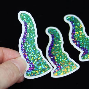 Könnte beinhalten: Drei regenbogenfarbene Glitzeraufkleber in Grün und Lila, die die Form eines gebogenen Horns haben. Die Aufkleber haben einen weißen Rand.