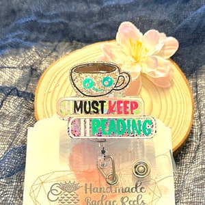 May include: A sparkly, glittery badge reel with a coffee cup, books, and the text "Must Keep Reading". The reel is attached to a clear plastic badge holder with a gold metal clip and a silver metal ring. The badge reel is on a wooden surface with a pink flower.