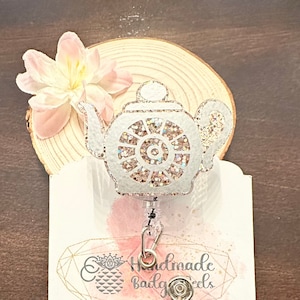 May include: A white glitter teapot badge reel with a pink and white flower and a wooden background. The reel has a clear plastic strap and a metal clip. The text on the card reads "Handmade Badge Reels" and "linktr.ee/craftternooncreating".