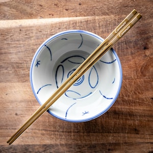 May include: A pair of gold chopsticks with the word "Connor" engraved on them, resting on a white bowl with a blue and white floral design.