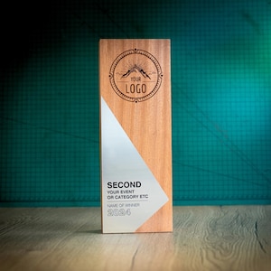 Puede incluir: Una placa de premio de madera con una incrustación de metal plateado. La placa tiene un logotipo de montaña con el texto "YOUR LOGO" y el texto "SECOND YOUR EVENT OR CATEGORY ETC NAME OF WINNER 2024".