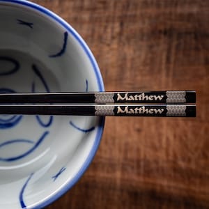 May include: A pair of black chopsticks with gold lettering that reads "Matthew" on each chopstick. The chopsticks are resting on the rim of a white bowl with a blue and white design.