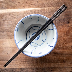 May include: A white ceramic bowl with a blue and white design, and a pair of black chopsticks with the name "Matthew" engraved on them.