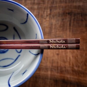 May include: A pair of rose gold chopsticks with the name "Nichola" engraved on each chopstick. The chopsticks are resting on a blue and white ceramic bowl.