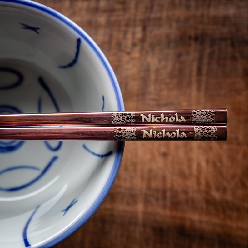 May include: A pair of rose gold chopsticks with the name "Nichola" engraved on each chopstick. The chopsticks are resting on a blue and white ceramic bowl.