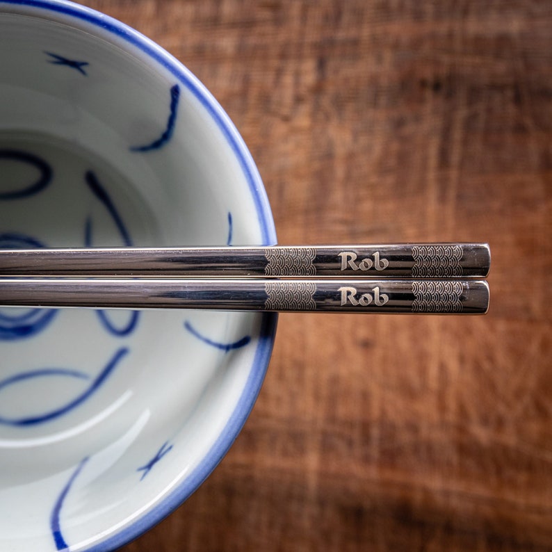 May include: A pair of silver chopsticks with the word "Rob" engraved on each stick. The chopsticks are resting on a white bowl with a blue and white floral design.