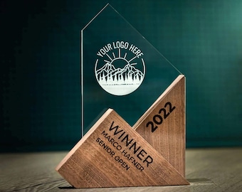 May include: A wooden and clear acrylic award trophy with the text "Winner 2022" and "Marco Hafner Senior Open". The top of the trophy features a mountain scene with a sun and the text "Your Logo Here".