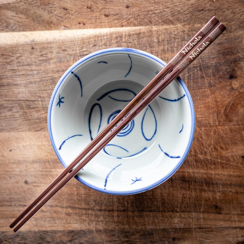 May include: A pair of rose gold chopsticks with the name "Nicholas" engraved on them, resting on a white bowl with blue and white floral designs.