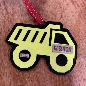 Può includere: Un ornamento in legno a forma di autocarro con cassone ribaltabile giallo con bordi neri. L'ornamento ha il nome "Kashton" e l'anno "2022". Un nastro rosso con cuciture dorate è attaccato per appenderlo.