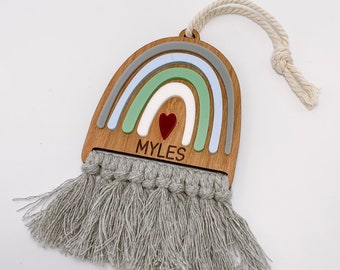 Könnte beinhalten: Wandbehang in Regenbogenform aus Holz mit Makramee-Fransen. Der Regenbogen hat Schichten aus Grau, Blau, Grün und Weiß, mit einem roten Herz und dem Namen "MYLES" in Schwarz. Ein cremefarbenes Seil ist zum Aufhängen befestigt.