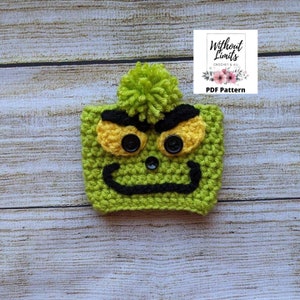 May include: Green crocheted coffee cozy with a Grinch face, black eyebrows, yellow eyes, and a black mouth. The cozy has a green pom-pom on top. The text "Without Limits Crochet & All PDF Pattern" is on a white tag with pink flowers.
