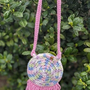 May include: A small, round crocheted bag with a pink strap and fringe. The bag is made with a variety of pastel colors, including pink, blue, green, and white.