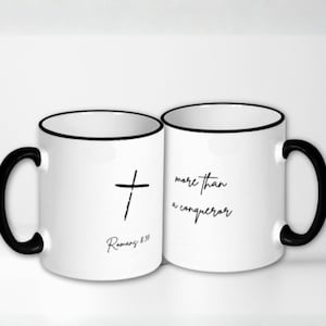 May include: Two white ceramic mugs with black handles and rims. One mug features a black cross and the text "Romans 8:37". The other mug has the phrase "more than a conqueror" in black script.