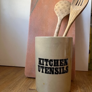 May include: A beige ceramic kitchen utensil holder with the words "KITCHEN UTENSILS" in black. Two wooden spoons are inside the holder, one with heart-shaped designs and the other with slots. A pink chopping board is in the background.