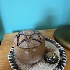 May include: A small, round, ceramic trinket box with a brown glaze and a blue and brown geometric design. The box is sitting on a white and brown cowhide coaster.
