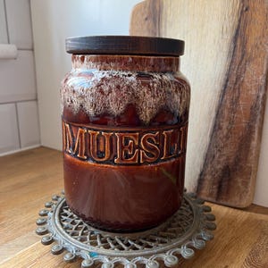 May include: A brown ceramic jar with a wooden lid, labelled "MUESLI" in embossed letters. The jar sits on a decorative metal trivet. The jar has a textured, dripped glaze effect in shades of brown and cream.