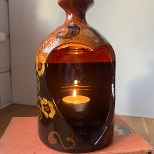 May include: A ceramic candle holder with a heart-shaped aperture, holding a lit tealight candle. The holder is brown with floral and swirl designs in yellow and black. It rests on a stack of vintage books.