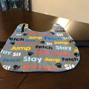 Op de afbeelding: Een grijs fleece honden trainingsvest met blauwe bies en zwarte banden. Het vest is bedrukt met de woorden "Jump Fetch Stay Roll Over Sit" in verschillende kleuren.