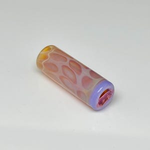 Könnte beinhalten: Eine zylindrische Glasperle mit einem durchscheinenden pfirsich- und orangefarbenen Wabenmuster auf der Oberfläche, abgeschlossen mit einem einfarbigen hellvioletten Band an einem Ende und einer hohlen Öffnung zum Auffädeln auf eine Schnur.