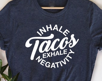 Könnte beinhalten: Marineblaues T-Shirt mit weißem Text "Inhale Tacos Exhale Negativity" in einem Kreis. Das T-Shirt hängt an einem Holzbügel. Grünzeug ist in der unteren linken Ecke zu sehen.
