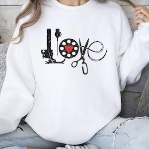 May include: White sweatshirt with a black graphic design that spells "LOVE" using a sewing machine, a spool of thread with a red heart, and scissors. The sweatshirt is a long-sleeved crew neck.