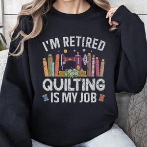 May include: Black sweatshirt with the text "I'M RETIRED QUILTING IS MY JOB" in white. The graphic features a sewing machine, spools of thread, and fabric rolls. The sweatshirt is a comfortable, casual garment.