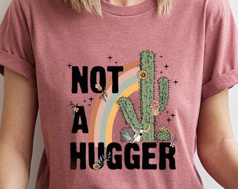 Puede incluir: Una camiseta de color malva claro con un gráfico de cactus y el texto "NOT A HUGGER".