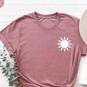 May include: A heather mauve t-shirt with a white sun graphic on the front. The shirt is laid flat on a white surface with a straw hat and a white mug.