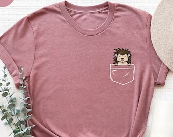 May include: A mauve-colored t-shirt with a pocket design featuring a cartoon hedgehog. The hedgehog is brown and smiling. A white hat and mug are visible in the background. The shirt is knotted at the bottom.