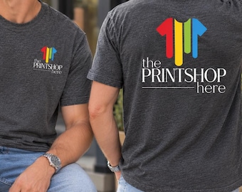 Puede incluir: Una camiseta de manga corta gris carbón con un logotipo pequeño en el pecho izquierdo y una versión grande en la espalda. El logotipo consta de cuatro barras verticales en rojo, amarillo, verde y azul, con el texto "the PRINTSHOP here" impreso debajo.