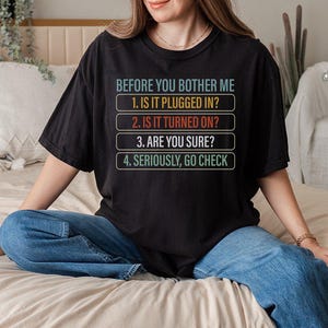 May include: Black t-shirt with the text "BEFORE YOU BOTHER ME" and a numbered checklist: "1. IS IT PLUGGED IN?", "2. IS IT TURNED ON?", "3. ARE YOU SURE?", and "4. SERIOUSLY, GO CHECK". The shirt is paired with blue jeans.