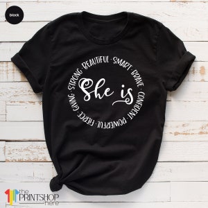 May include: Black t-shirt with a white circle graphic that says "She is" surrounded by the words "powerful, fierce, giving, strong, beautiful, smart, brave, confident".