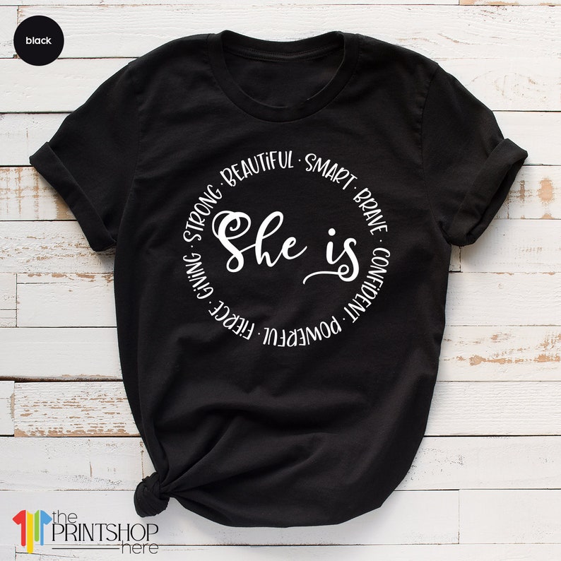 May include: Black t-shirt with a white circle graphic that says "She is" surrounded by the words "powerful, fierce, giving, strong, beautiful, smart, brave, confident".