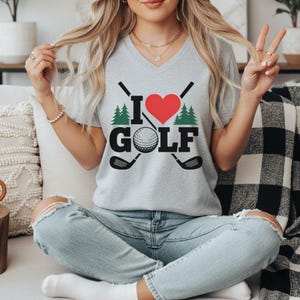 May include: A light grey V-neck t-shirt featuring the text "I ❤️ GOLF". The design includes a red heart, a golf ball, crossed golf clubs, and green pine trees. The shirt is made of a soft material.