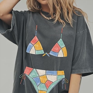 May include: A charcoal gray oversized t-shirt featuring a graphic print of a patchwork bikini. The bikini design consists of multi-colored geometric segments in shades of blue, yellow, red, and green, outlined with black stitching details.