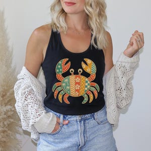 May include: A black tank top featuring a large, central graphic of a crab. The crab is composed of vertical, multi-colored patterned sections in shades of orange, green, and yellow, including palm tree, floral, and geometric motifs.