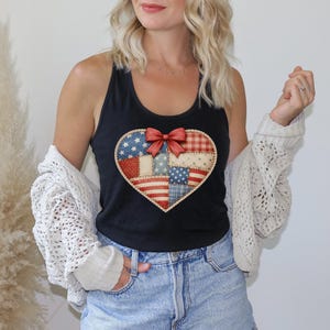Könnte beinhalten: Ein schwarzes Tanktop mit einer großen herzförmigen Grafik auf der Vorderseite. Das Herz besteht aus einem Patchwork-Design in Rot, Weiß und Blau mit Sternen- und Streifenmustern und ist in der Mitte mit einer auffälligen roten Schleife verziert.