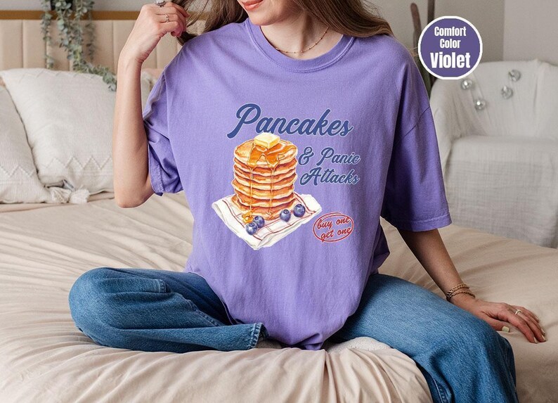 a woman sitting on a bed with a stack of pancakes on her shirt