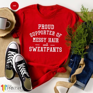 May include: Red t-shirt with white text that reads "Proud Supporter of Messy Hair and Sweatpants".