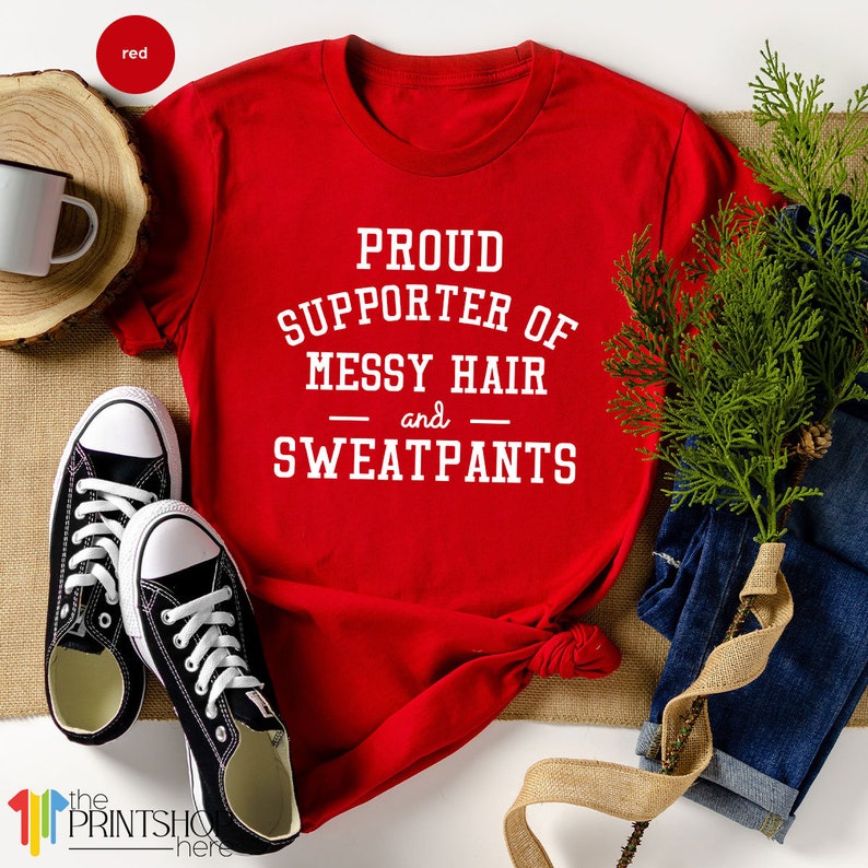May include: Red t-shirt with white text that reads "Proud Supporter of Messy Hair and Sweatpants".