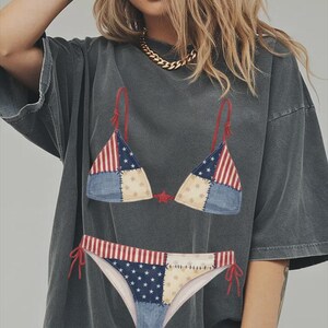May include: A charcoal gray oversized t-shirt featuring a graphic print of a two-piece bikini with a patchwork design. The bikini pattern incorporates red and white stripes, blue sections with white stars, and cream-colored fabric with star motifs.