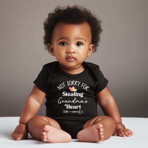 May include: A black baby bodysuit with the text "NOT SORRY FOR Stealing Grandma's Heart" in white script. The design includes two small hearts and an arrow. The baby is sitting on a white surface.