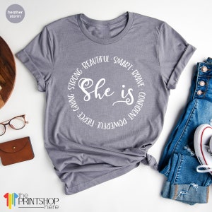 May include: Gray t-shirt with a white circle design that says "She is" with the words "Strong, Beautiful, Smart, Brave, Confident, Powerful, Fierce, Giving" surrounding the text.