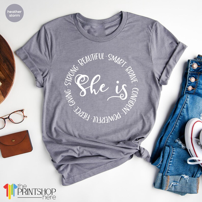 May include: Gray t-shirt with a white circle design that says "She is" with the words "Strong, Beautiful, Smart, Brave, Confident, Powerful, Fierce, Giving" surrounding the text.